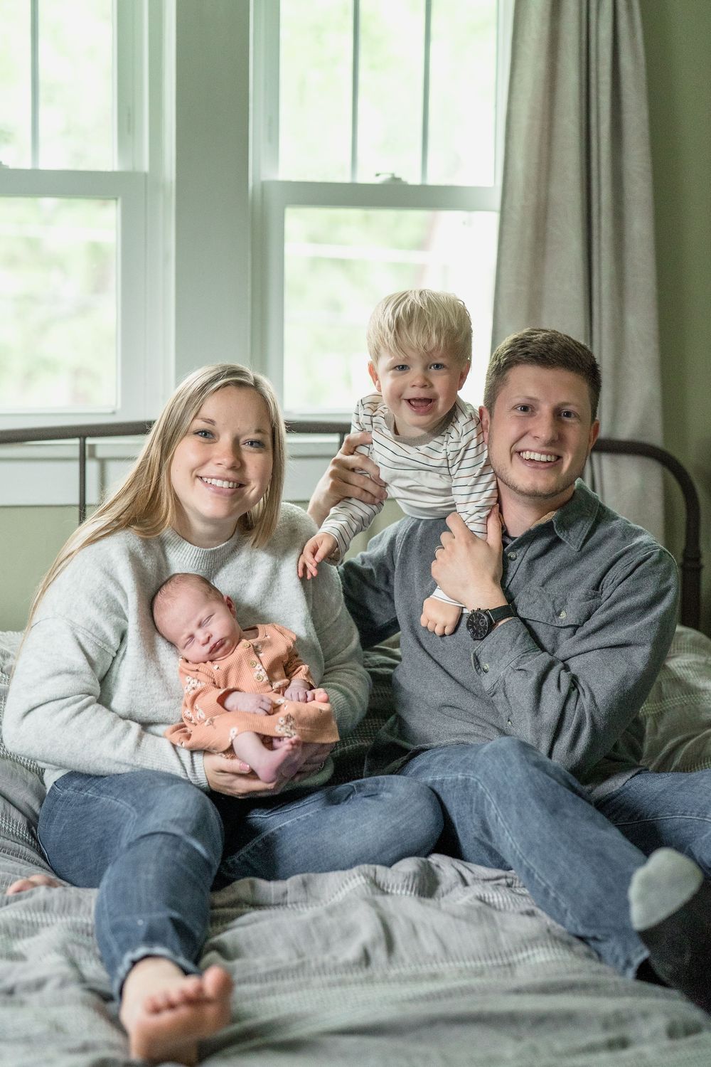 A family relaxes together on a bed near a window, sharing happy moments with their newborn baby and toddler in Cuyahoga Falls, Ohio Portrait