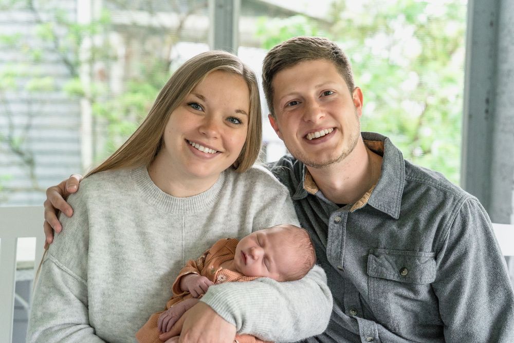 A couple sitting together on a couch hold their newborn baby while smiling at the camera in Cuyahoga Falls, Ohio Newborn Photo Session