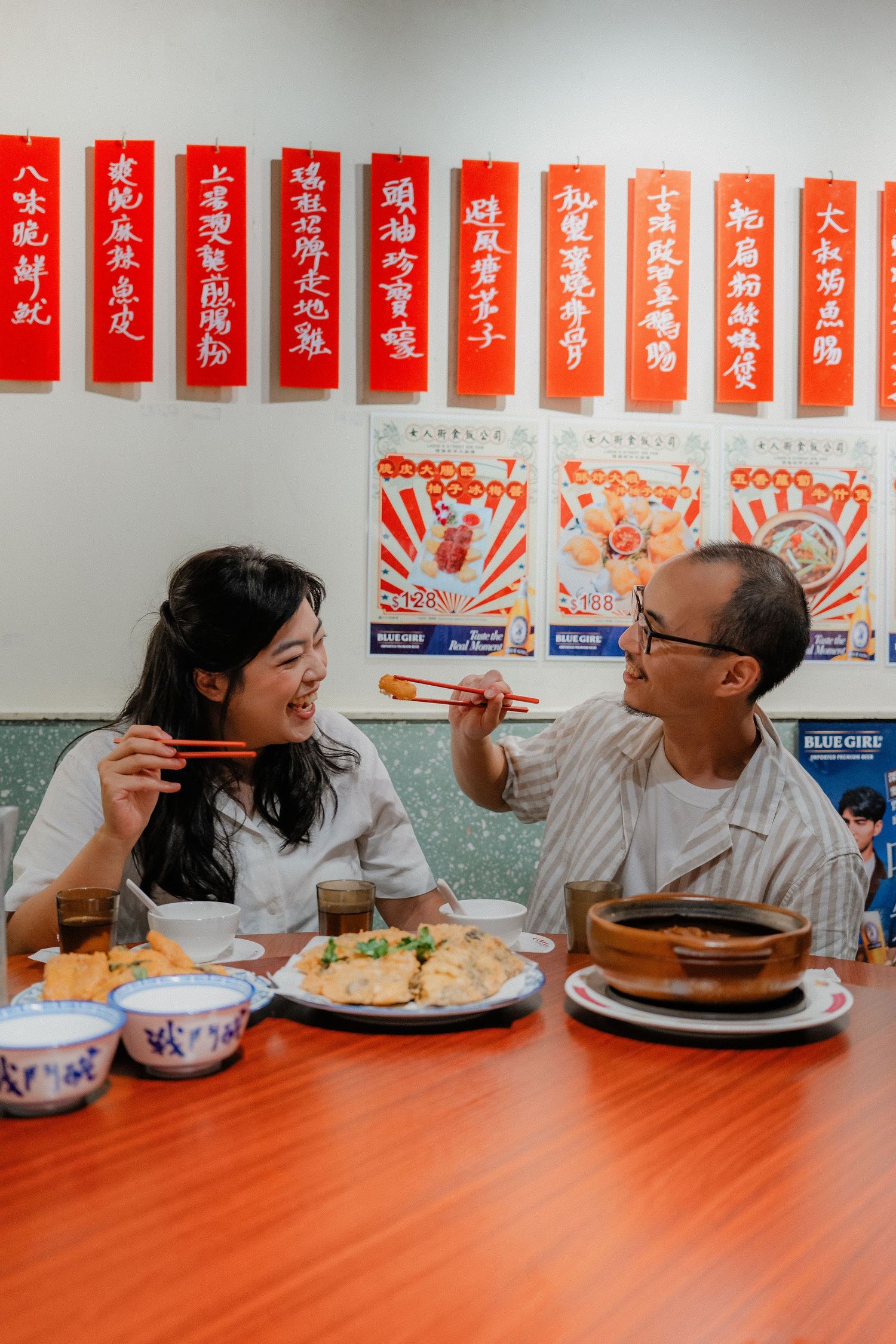 Two people enjoying a meal at a Chinese restaurant with traditional red banners and posters on the wall behind them.