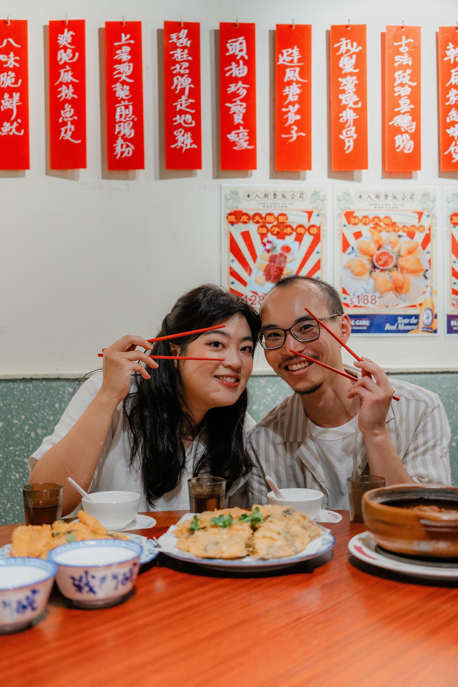 Two diners playfully posing with chopsticks at a Chinese restaurant under red decorative banners.