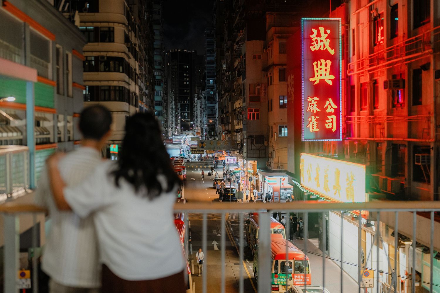 A couple overlooking a vibrant Hong Kong street at night with neon signs.