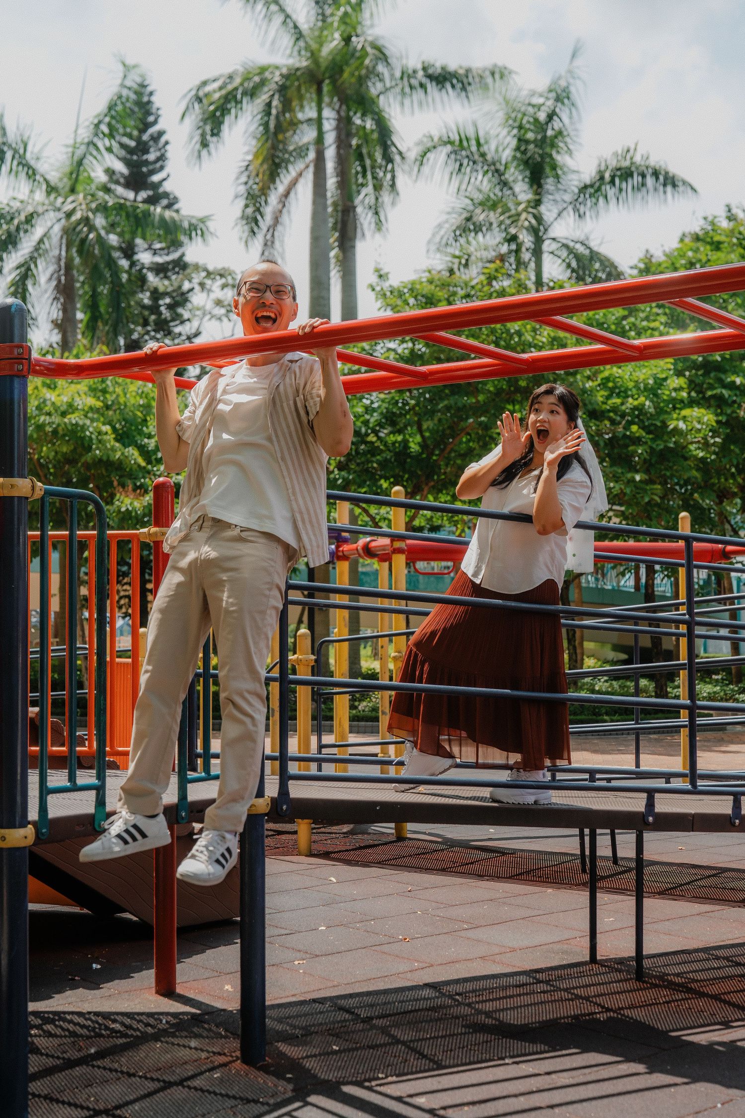 Two people play on outdoor exercise equipment in a park with tall palm trees in the background.
