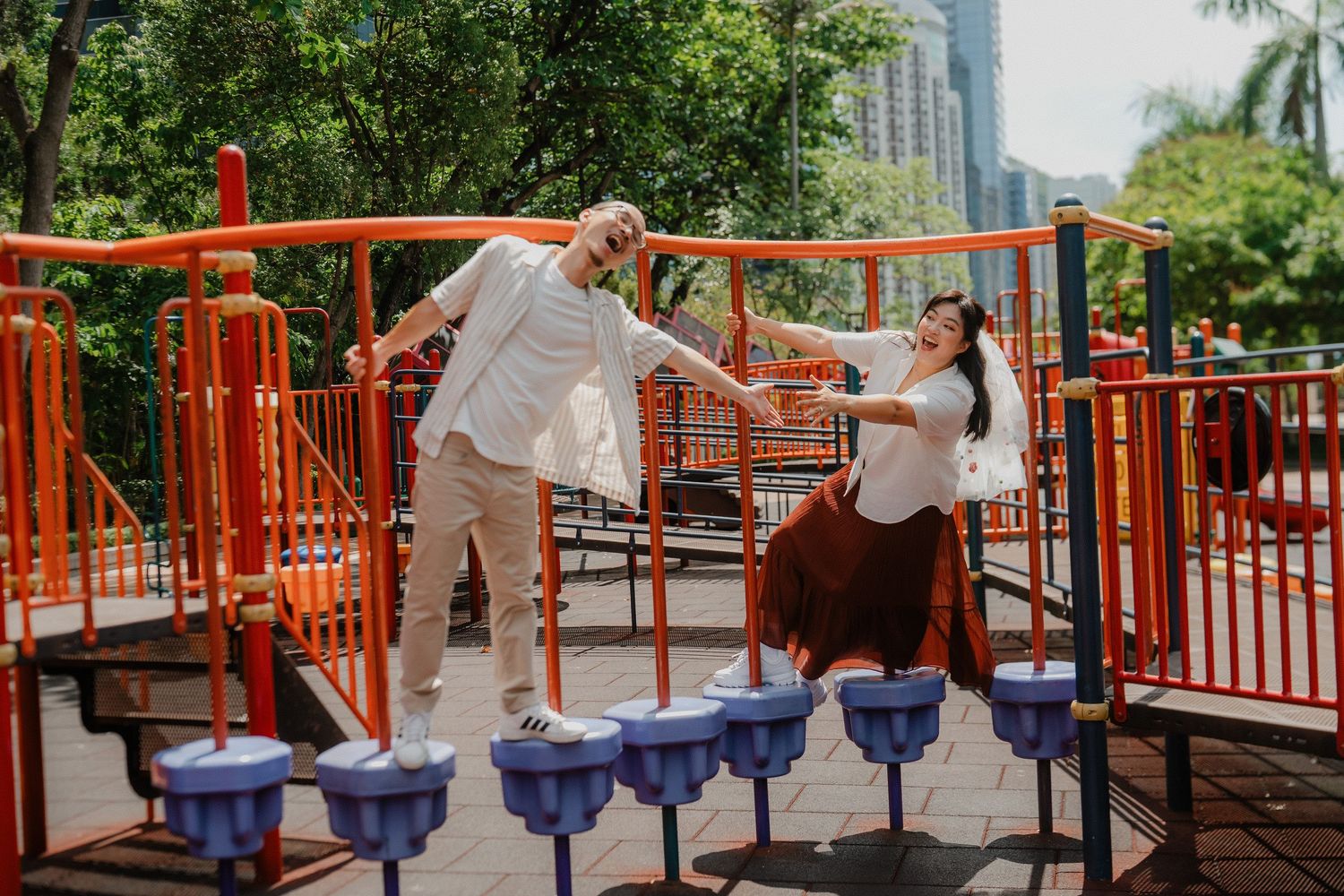 Couple enjoying a playful moment on a bright orange playground structure with blue seats in an urban park setting.