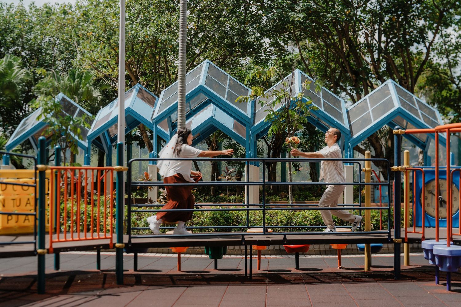 Colorful playhouses and playground equipment arranged in a row under shady trees at an outdoor park.