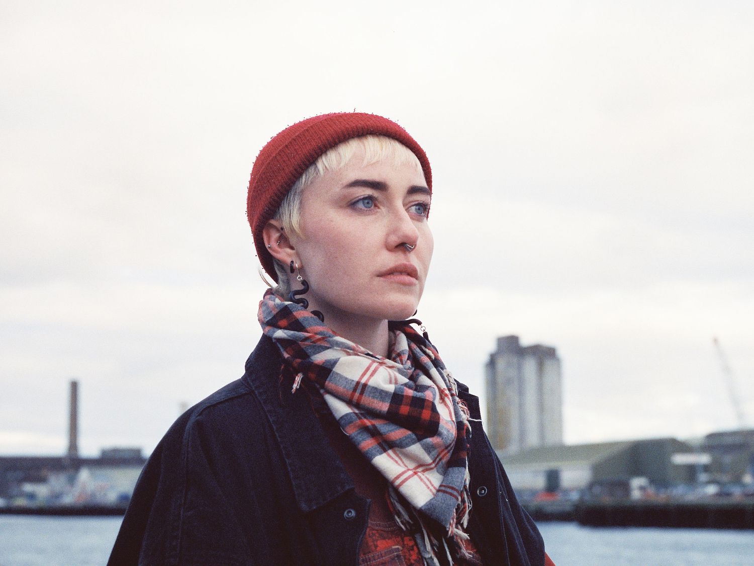 A person in a red beanie and plaid scarf looks out thoughtfully across a harbor on an overcast day.