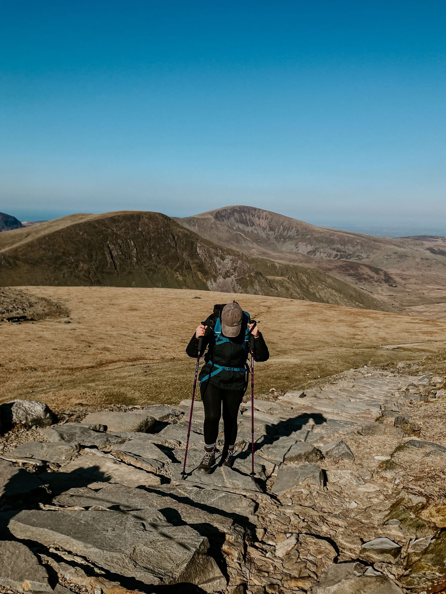 a photo of a lady in hiking clothes walking up snowdon mountain path. she is wearing a cap and walking with hiking stick.