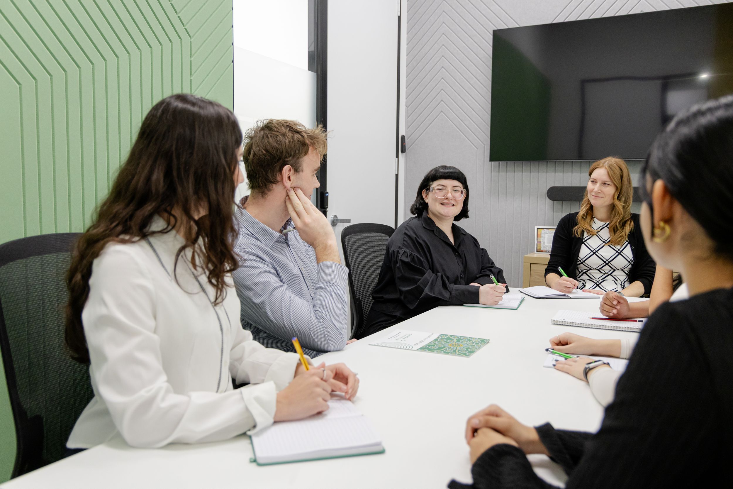 Business professionals gather around a conference table in a modern office space with green wall panels for a meeting.