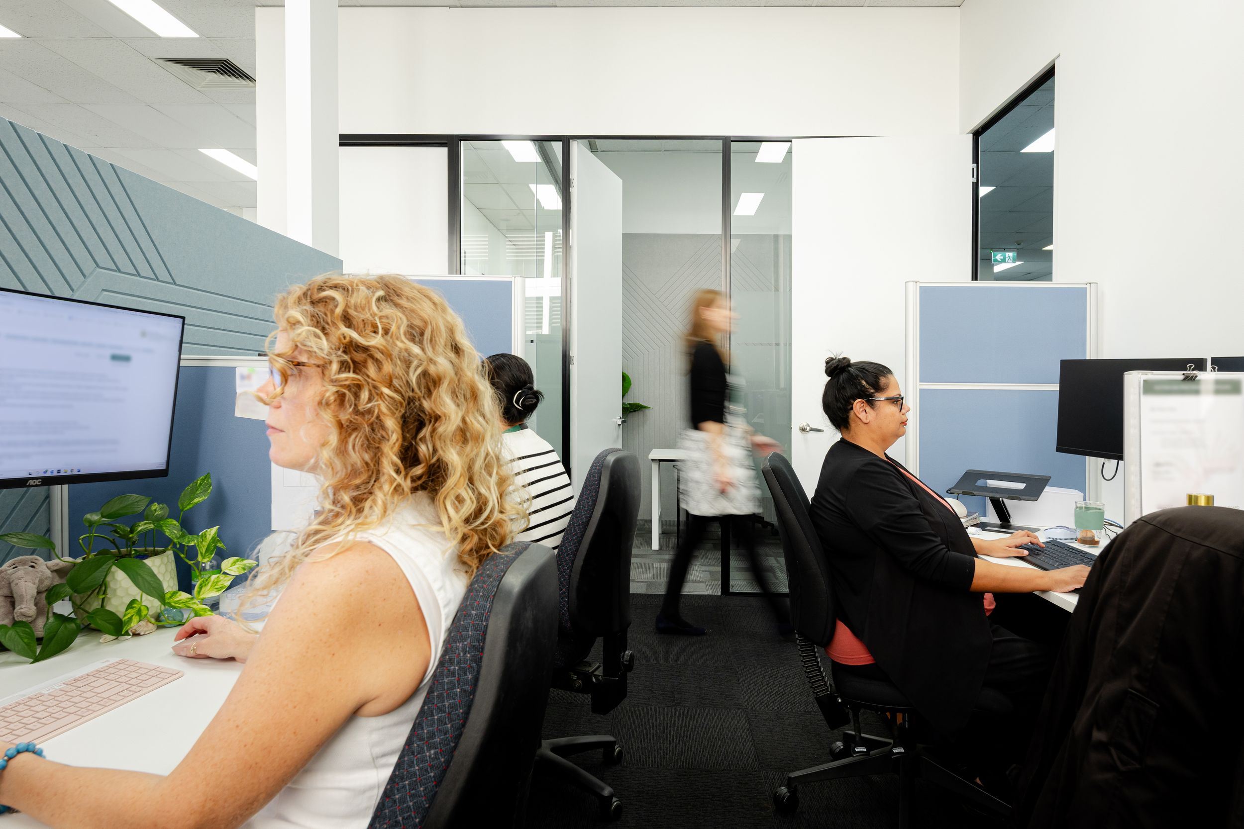 Office workers at their desks in a modern workspace with glass partitions and computer monitors.