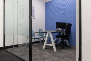 Modern office room with a glass desk, ergonomic chair, and bold blue accent wall against gray carpeted flooring.
