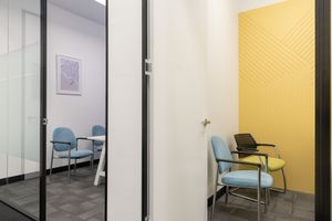 Modern office interior with glass partition, light blue chairs, and vibrant yellow accent wall in corner meeting space.