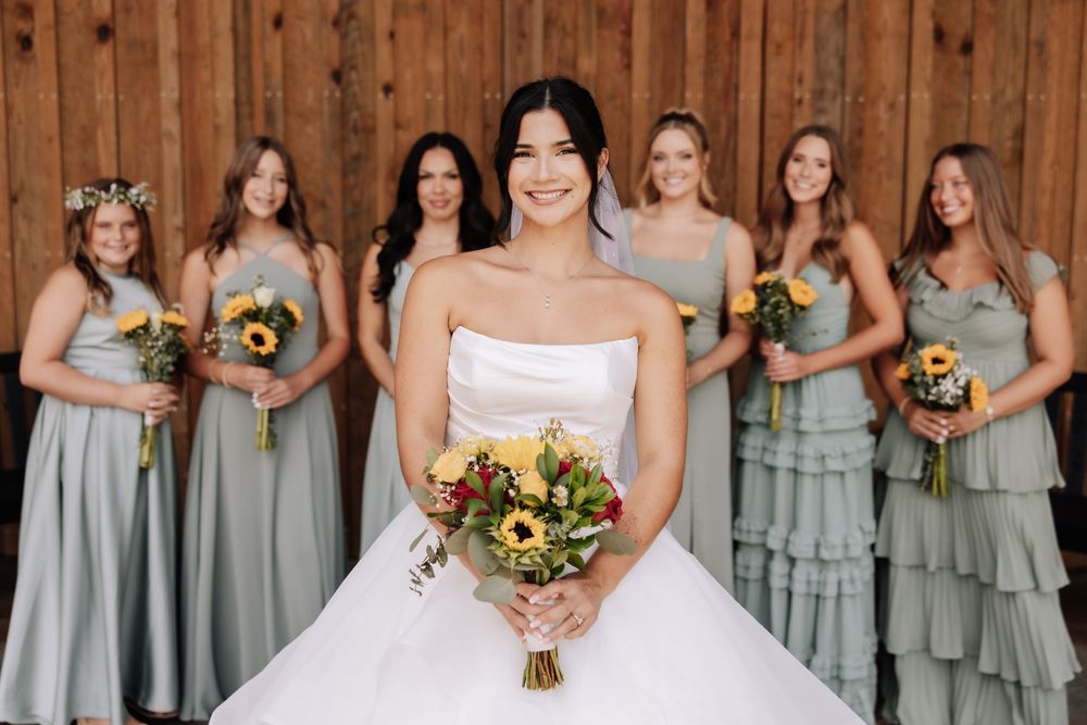A bridal party in soft sage green bridesmaid dresses stands together holding yellow sunflower bouquets.