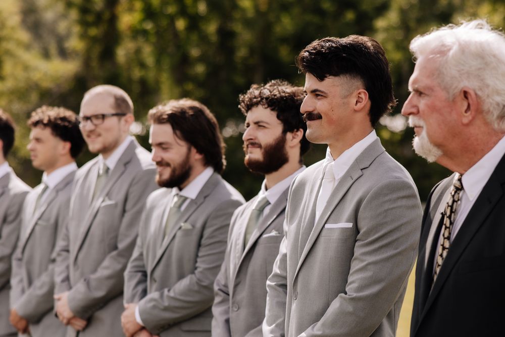 A row of groomsmen in matching light gray suits stand together at an outdoor wedding ceremony.