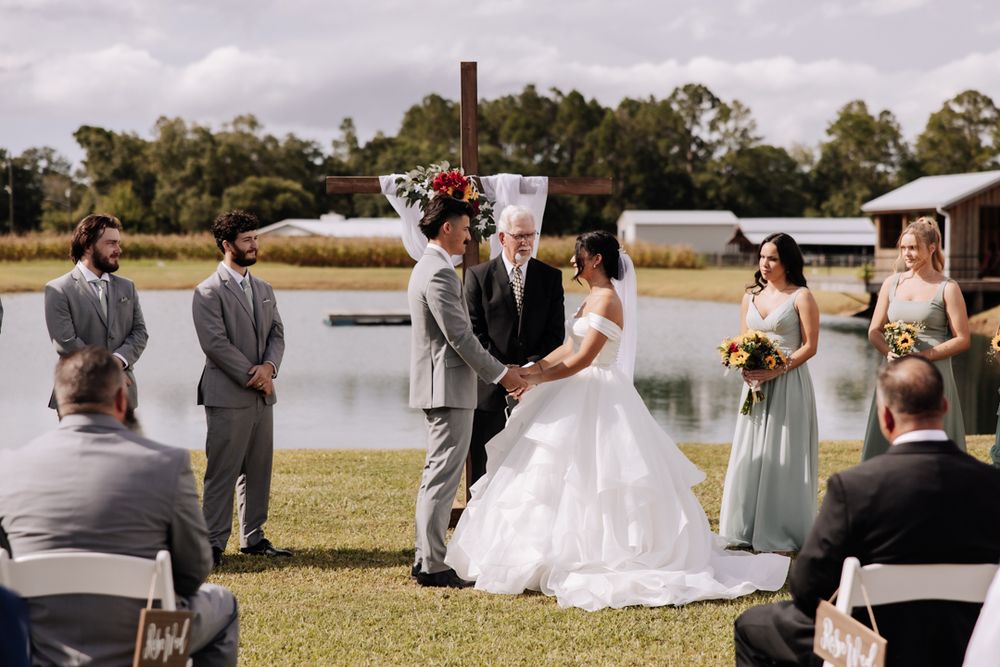 A scenic outdoor wedding ceremony takes place near a serene lake with guests seated in white chairs under cloudy skies.