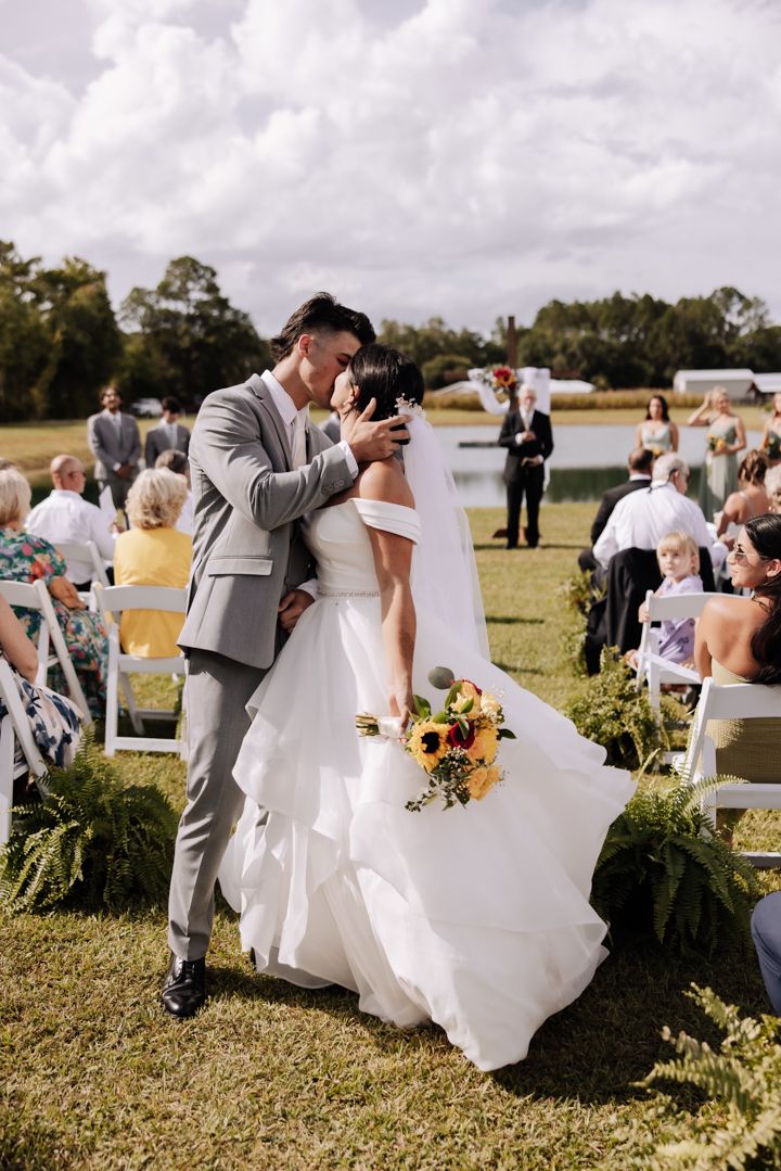 Outdoor wedding celebration scene with wedding party posing in a grassy field under dramatic cloudy skies.