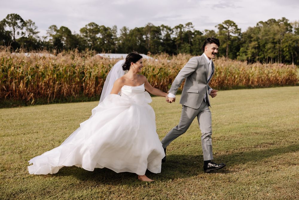 A couple in wedding attire runs playfully across a grassy field next to a cornfield on their special day.