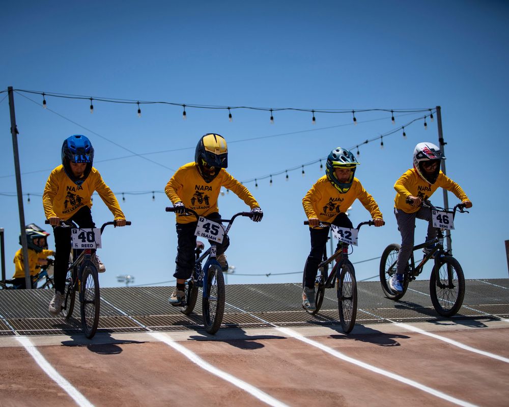 BMX rider performs a sequence of jumping tricks on a dirt track against a bright blue sky.