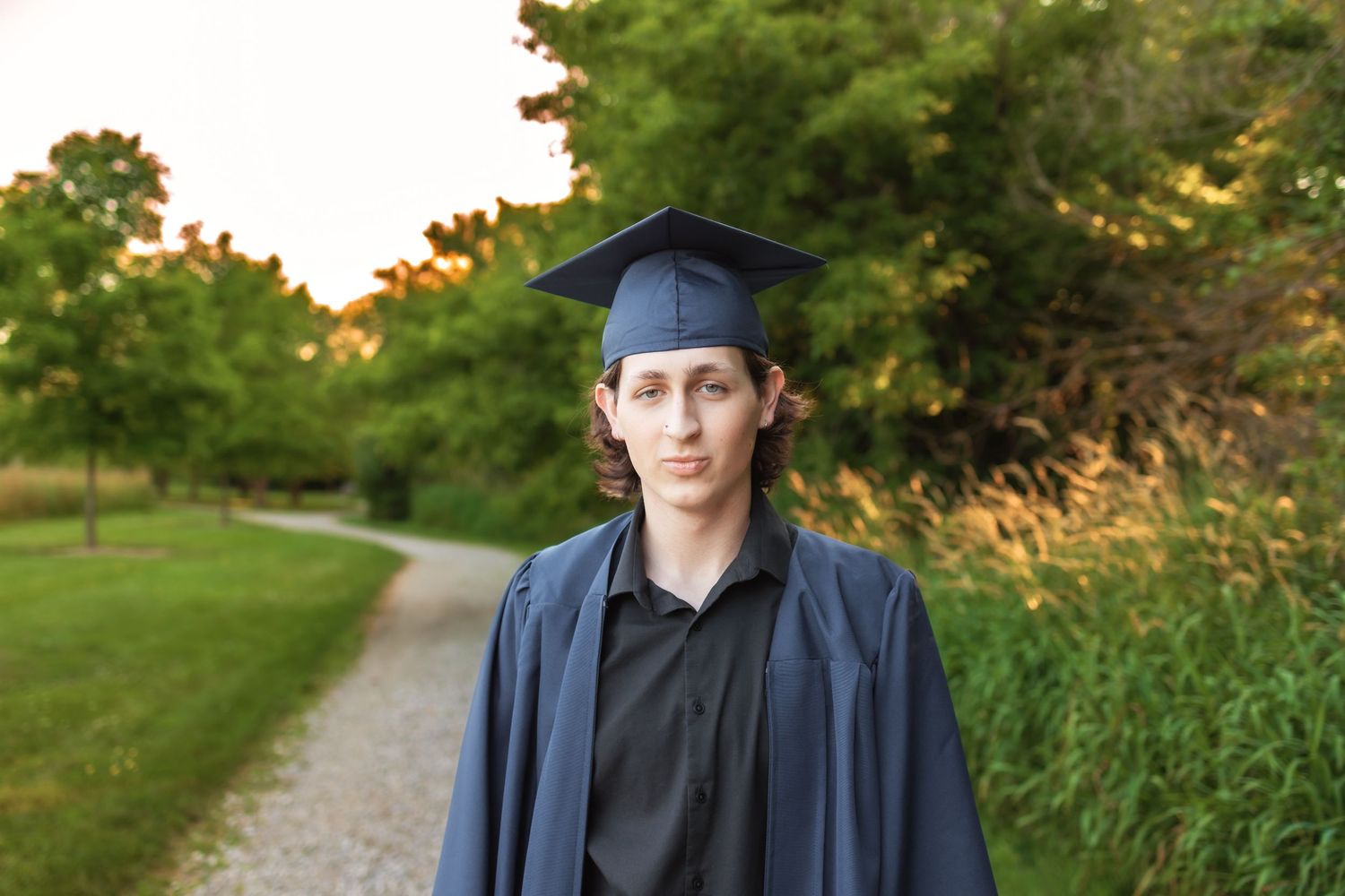 Graduate in cap and gown stands on winding path with trees and sunset light in background. Novi Michigan Senior Photographer