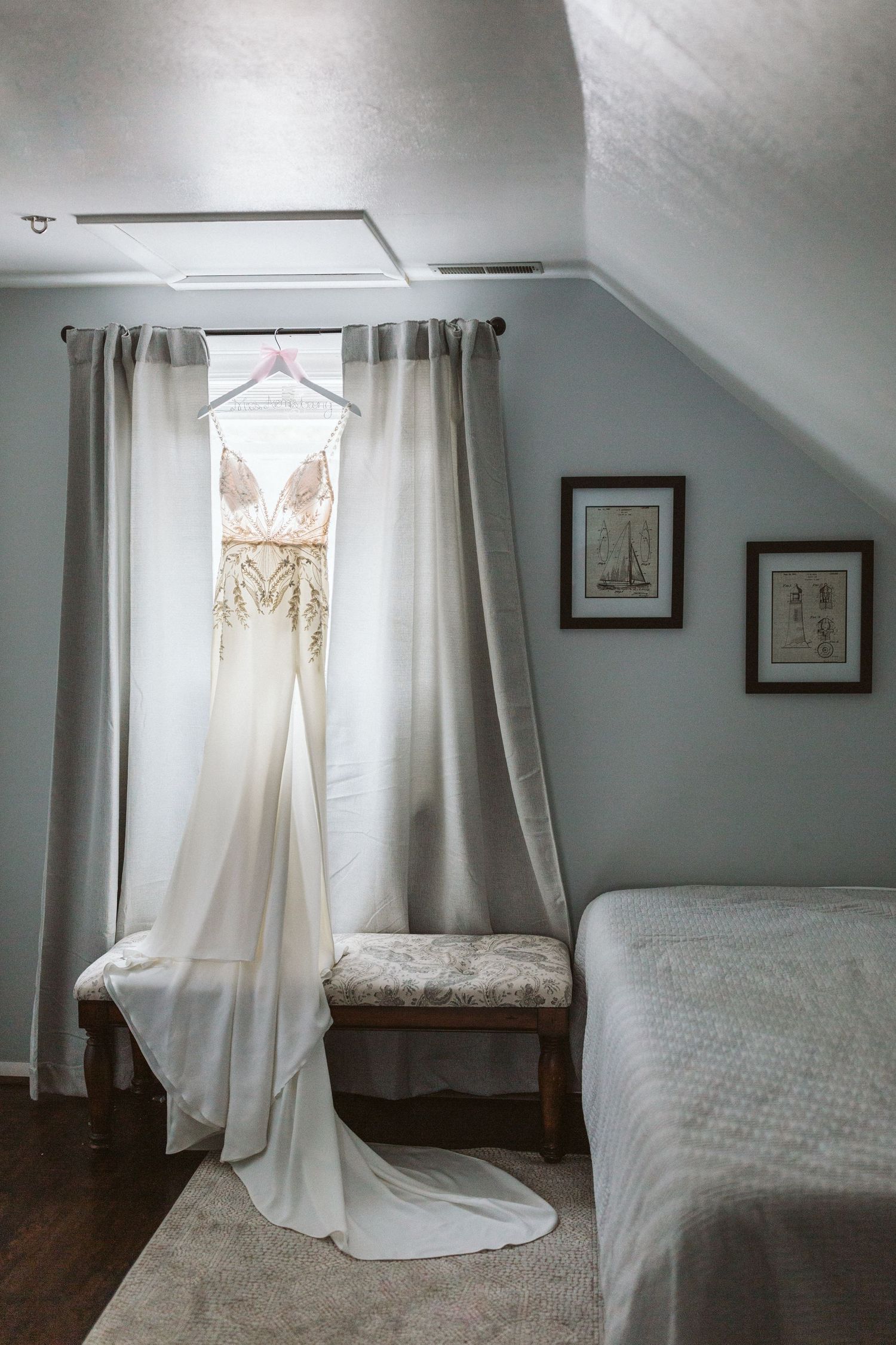 Elegant white wedding dress hanging in a soft-lit bedroom with gray walls and vintage decor.