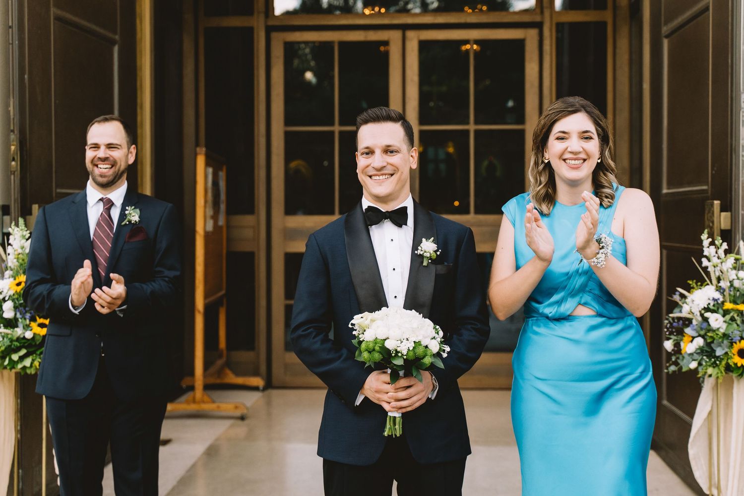 Wedding party members in formal attire share a joyful moment during a ceremony with decorative white floral arrangements.