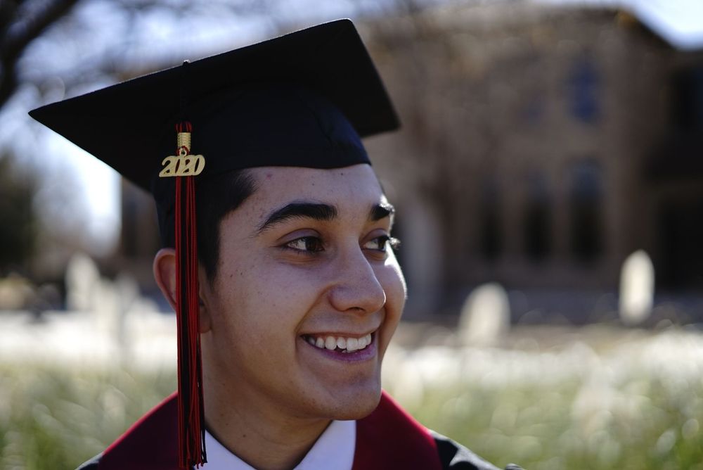 Graduate in red and black regalia smiles while wearing a mortarboard cap against a blurred building background. alt="Professional photograph