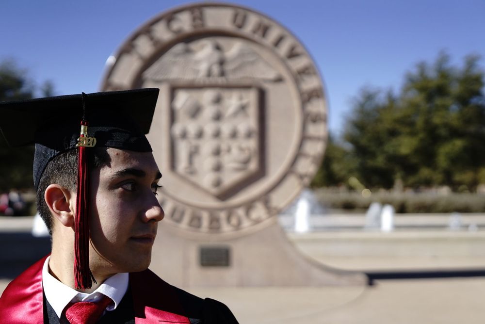 Graduate in red gown stands beside a large university seal monument on campus.  alt="Professional grad photography in Lubbock TX by H & Co.