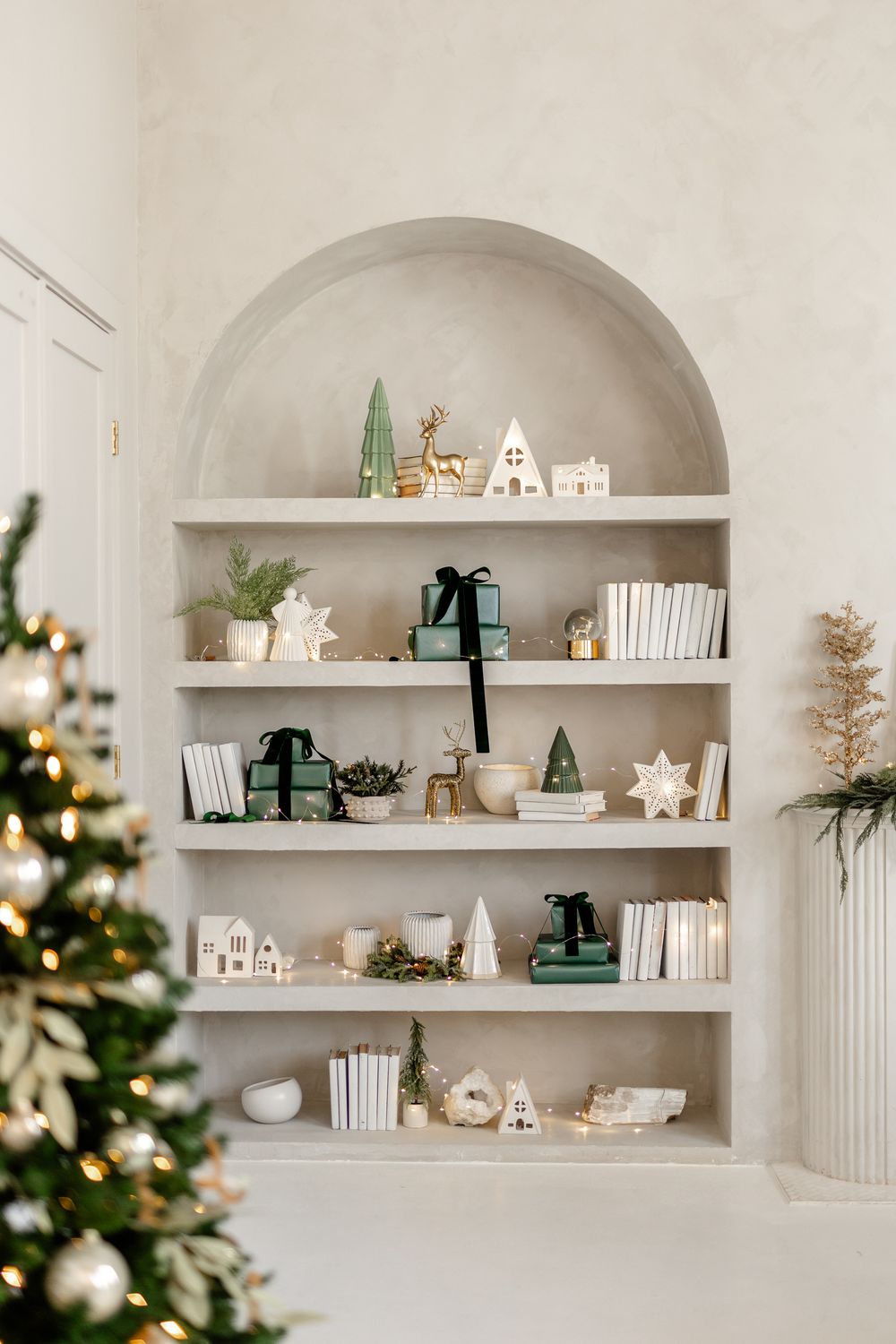 White built-in arched bookshelf decorated with holiday ornaments, green trees and white decorative houses near Christmas tree.