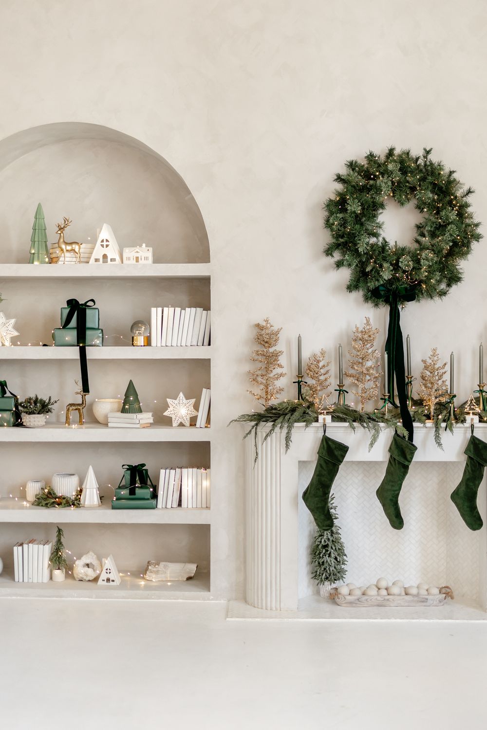 Minimalist Christmas decor with built-in shelves, green stockings, and evergreen wreath on white wall with holiday ornaments.