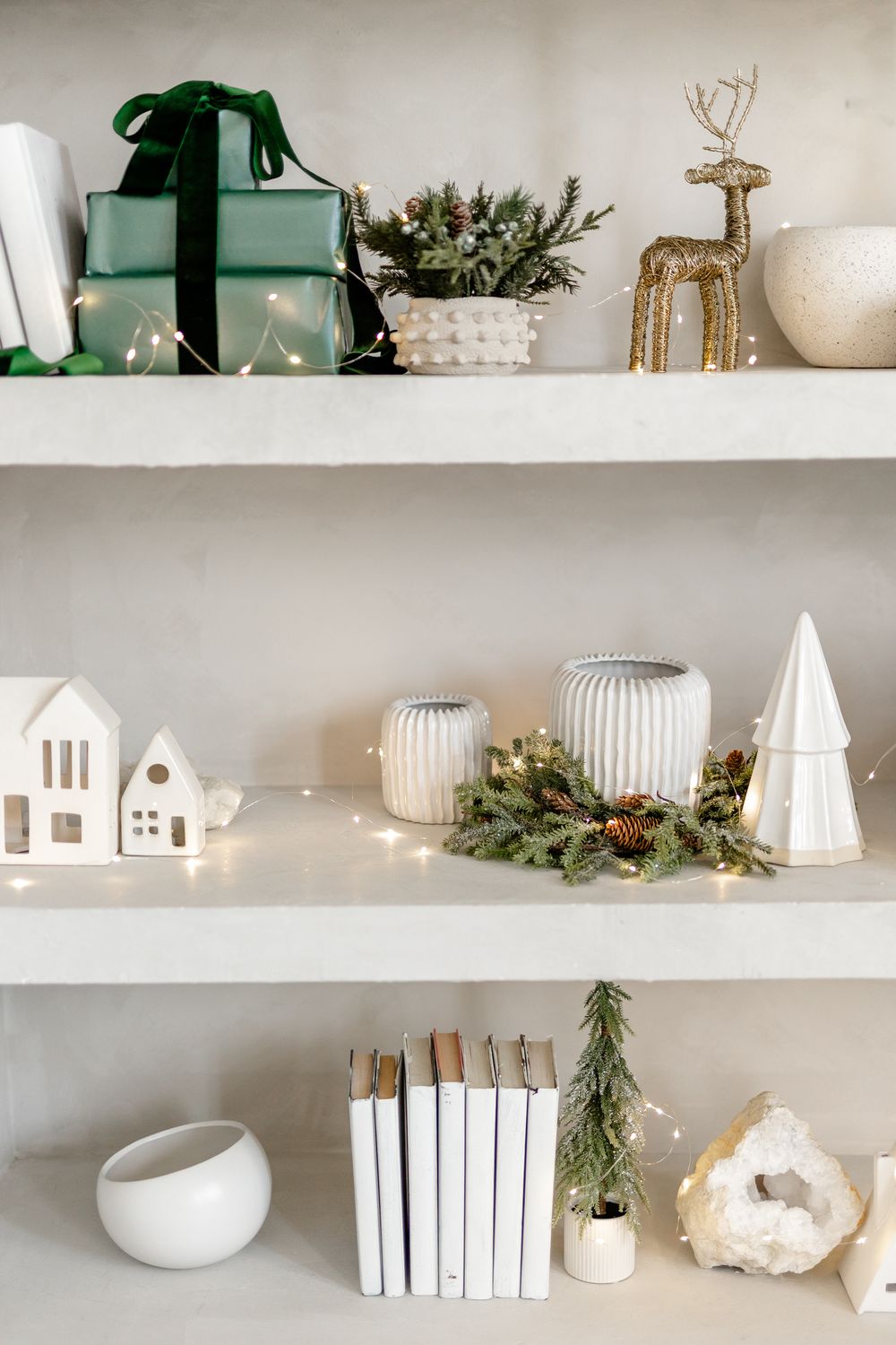 White ceramic houses and decorative objects arranged on minimalist floating shelves with green pine garland accents.