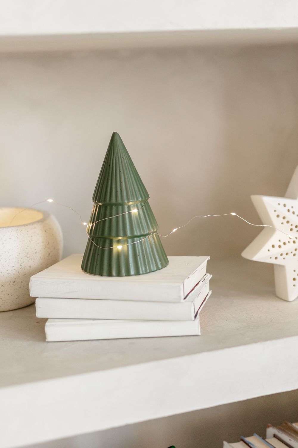 A green ceramic Christmas tree decoration sits atop stacked white books on a light surface.