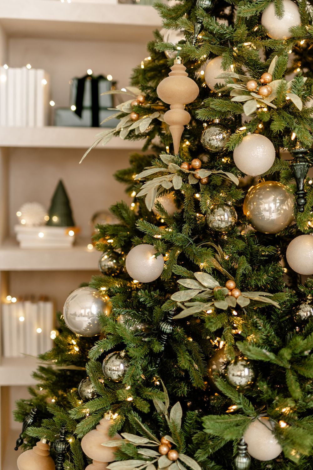 Elegant Christmas tree decorated with white and silver ornaments, green foliage, and warm lights against built-in shelving.