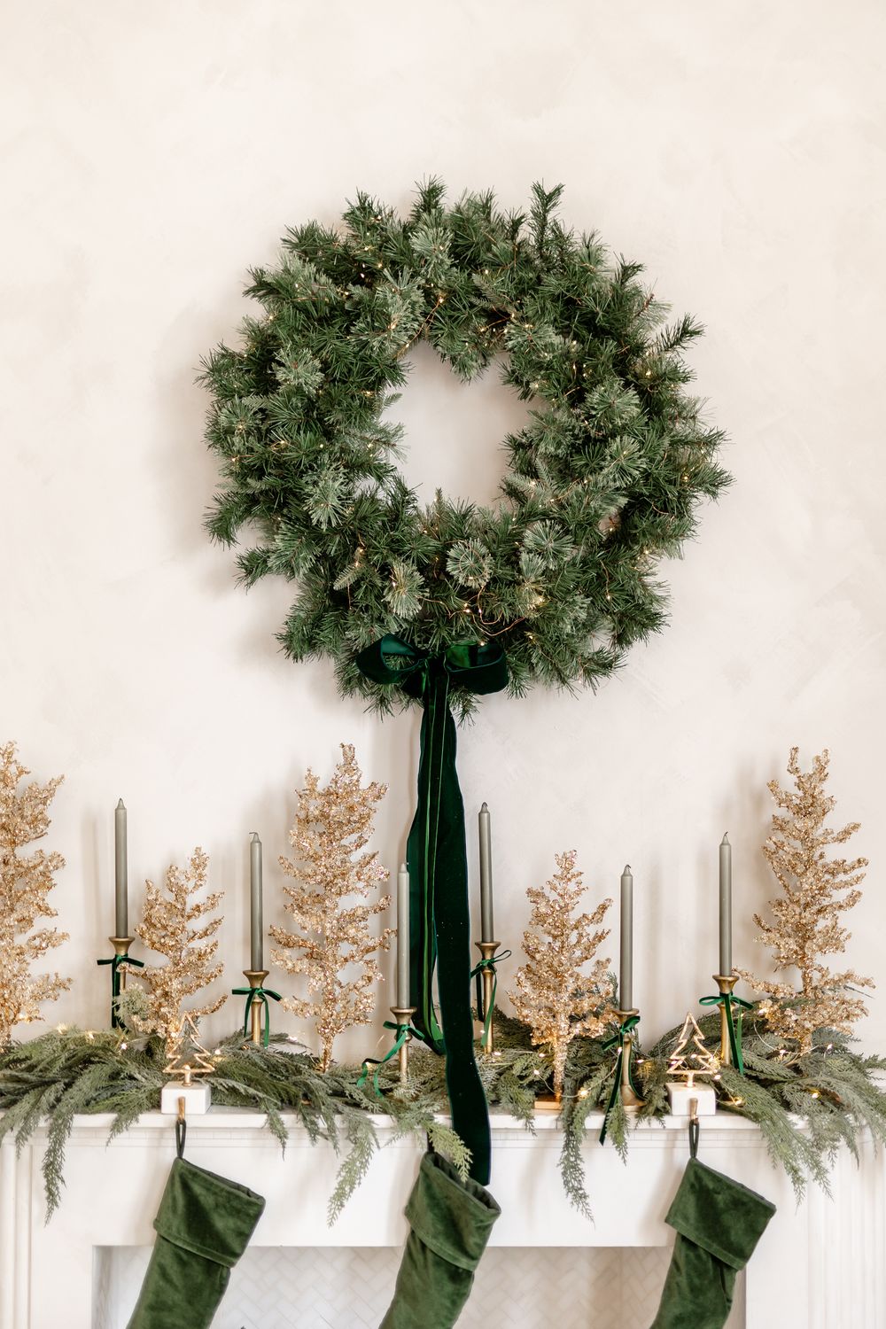Christmas mantel display with evergreen wreath, silver candles, gold miniature trees, and hanging green stockings.