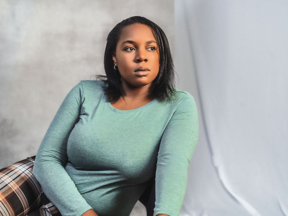 Person wearing a sage green long-sleeved top sits in contemplative pose against light background.