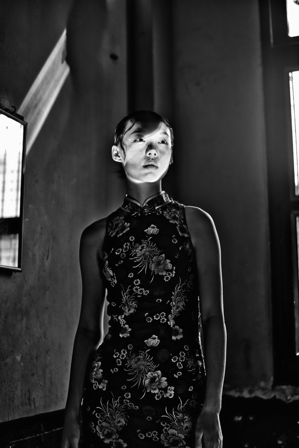 Dramatic black and white photograph of someone wearing a traditional Chinese qipao dress with floral embroidery in dark setting.