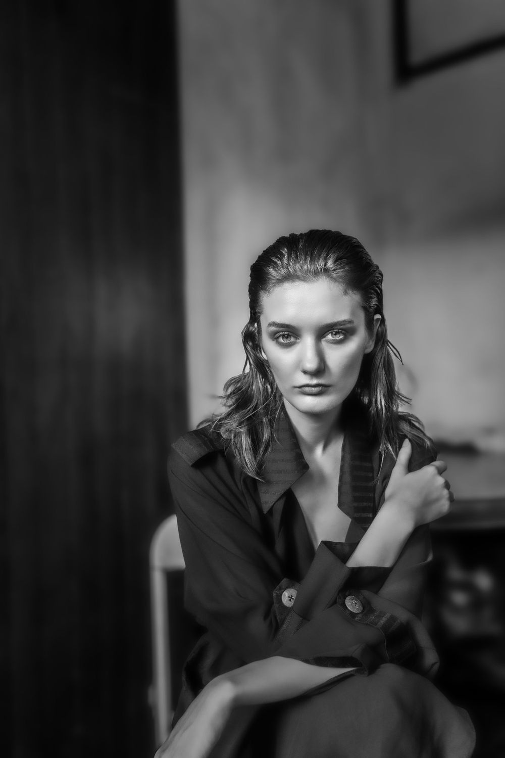 A dramatic black and white portrait of a model with wet hair wearing a dark blazer in a moody indoor setting.
