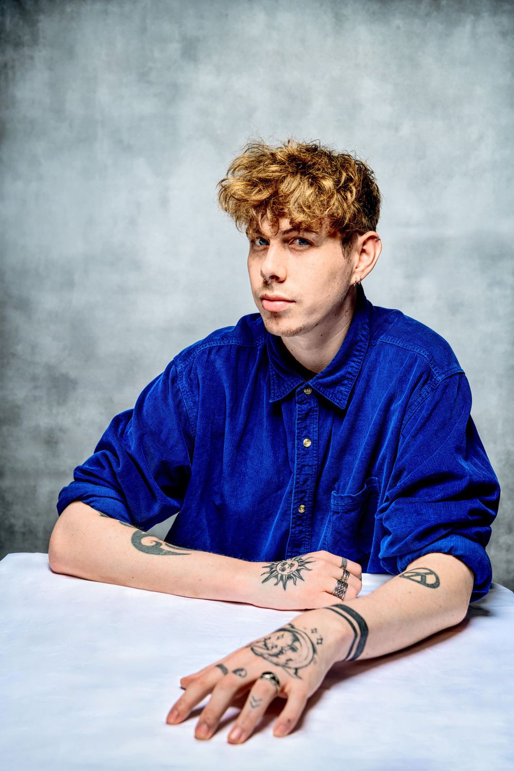 Portrait of someone in vibrant blue button-down shirt with curly blonde hair and arm tattoos against grey backdrop.