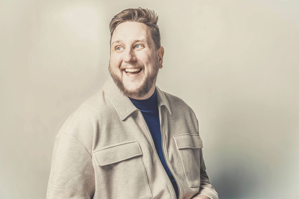 Professional portrait photo with someone wearing a beige jacket and blue sweater against a light neutral background.