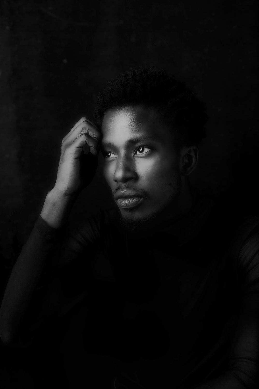 Dramatic black and white portrait with one hand raised to head, highlighting striking features against dark background.