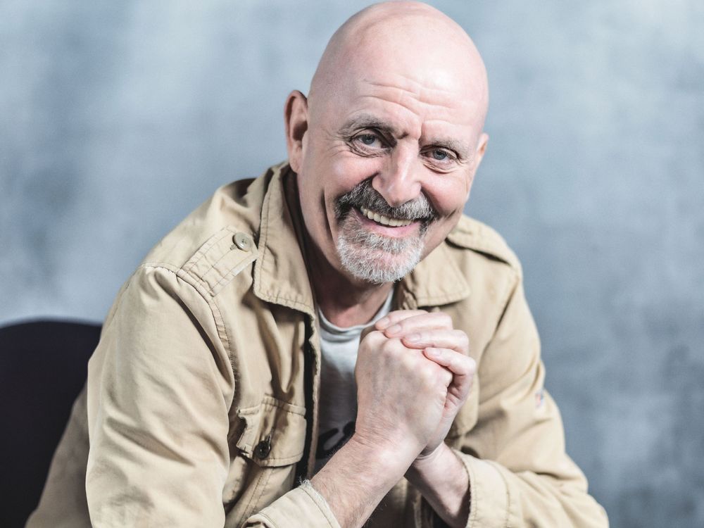 A smiling man in a beige jacket clasps his hands together while posing against a gray background.