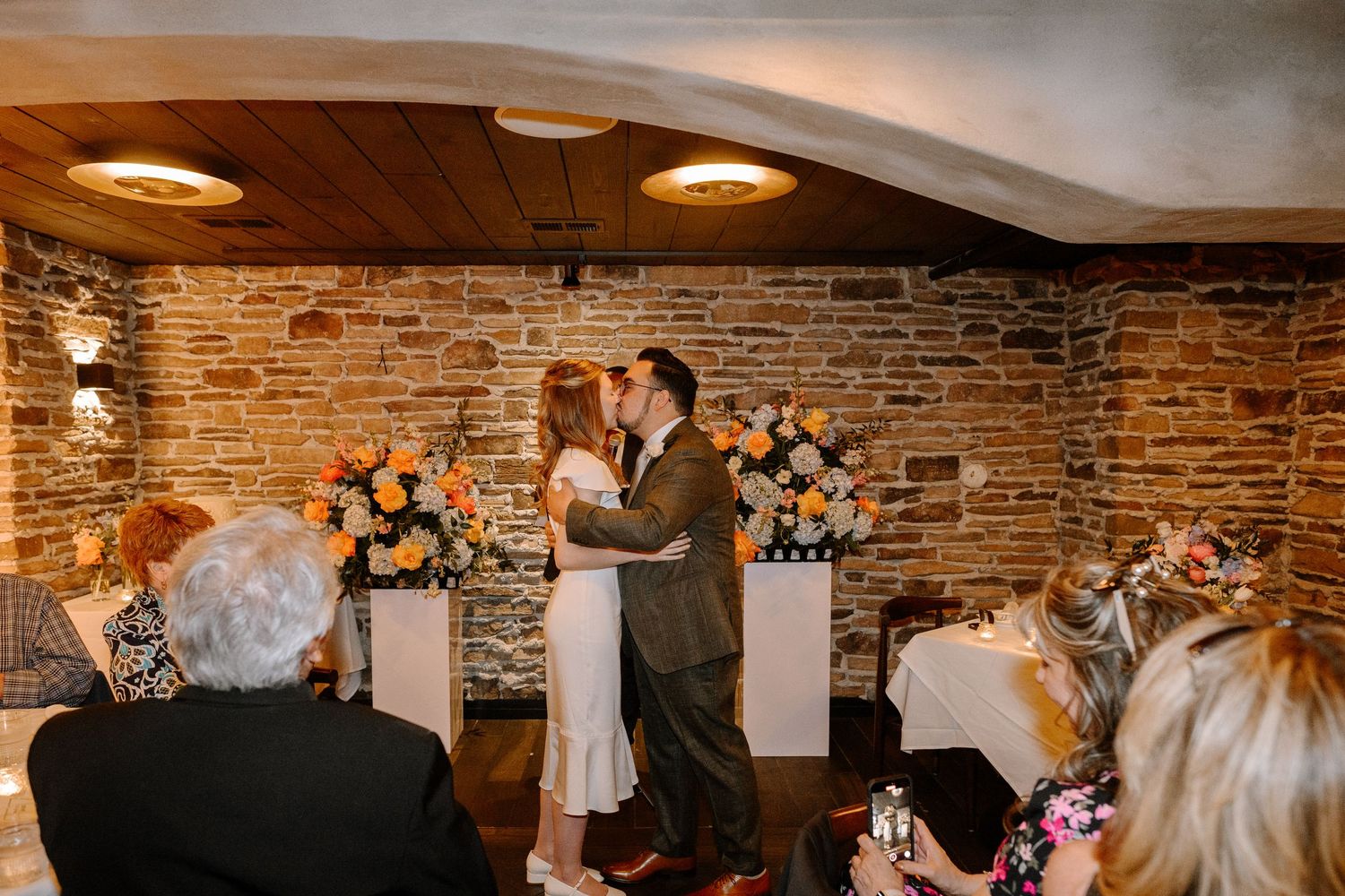 Wedding reception guests dancing and celebrating on a crowded dance floor in a rustic venue with exposed brick walls.