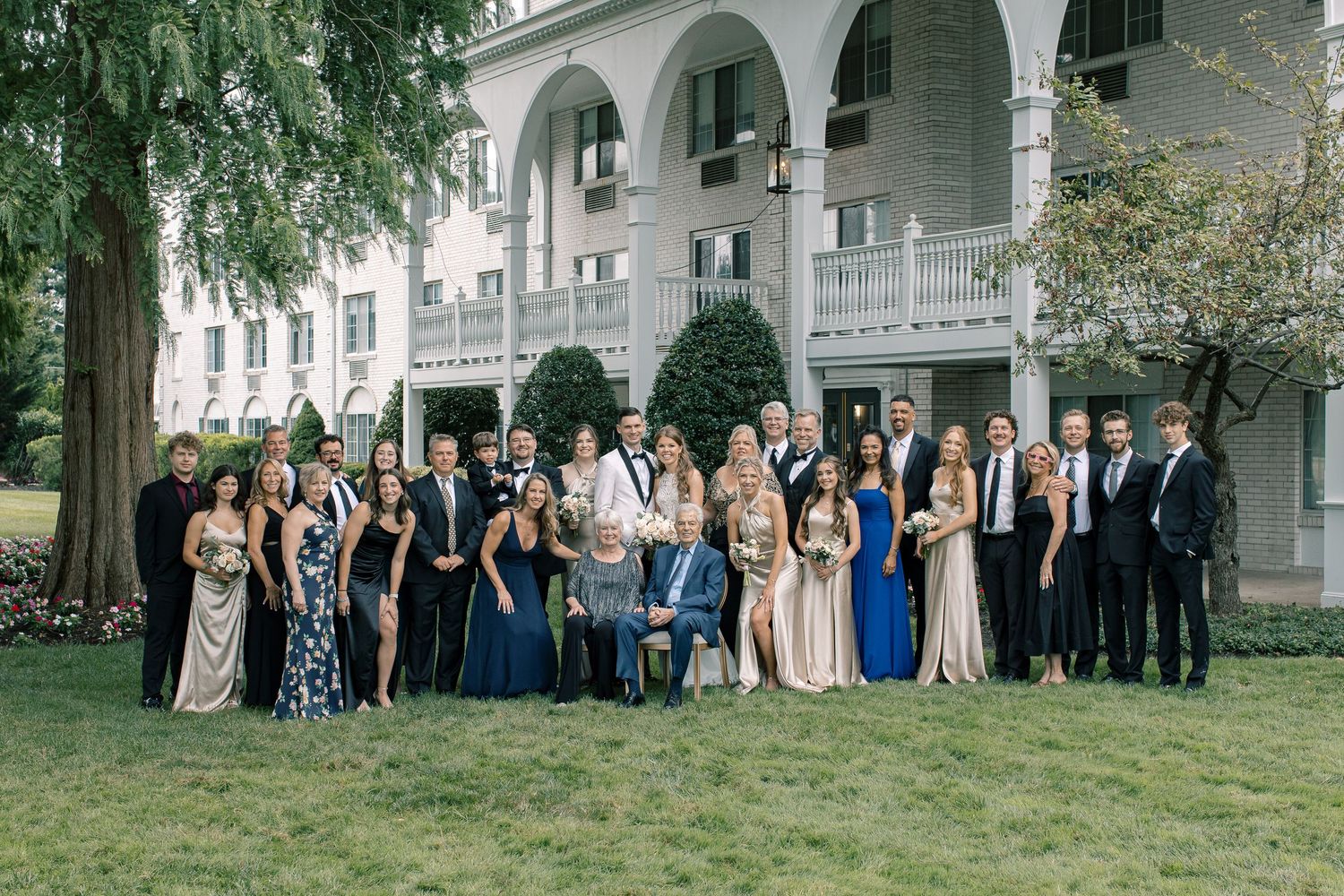 Large wedding party poses outdoors in formal attire beneath trees and arched columns of a building.