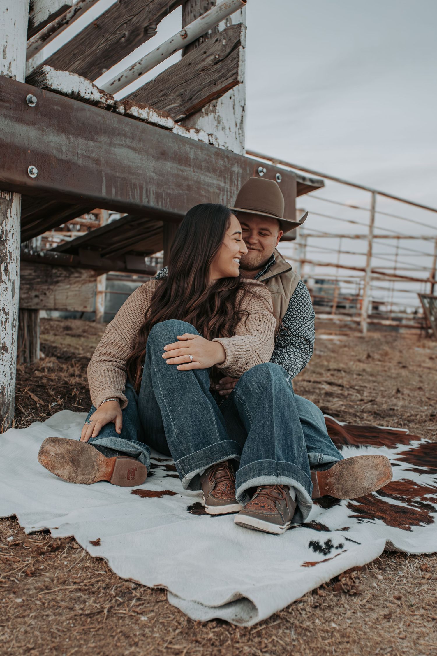A cozy couple sits together on a blanket in the snow near a rustic wooden structure at sunset.