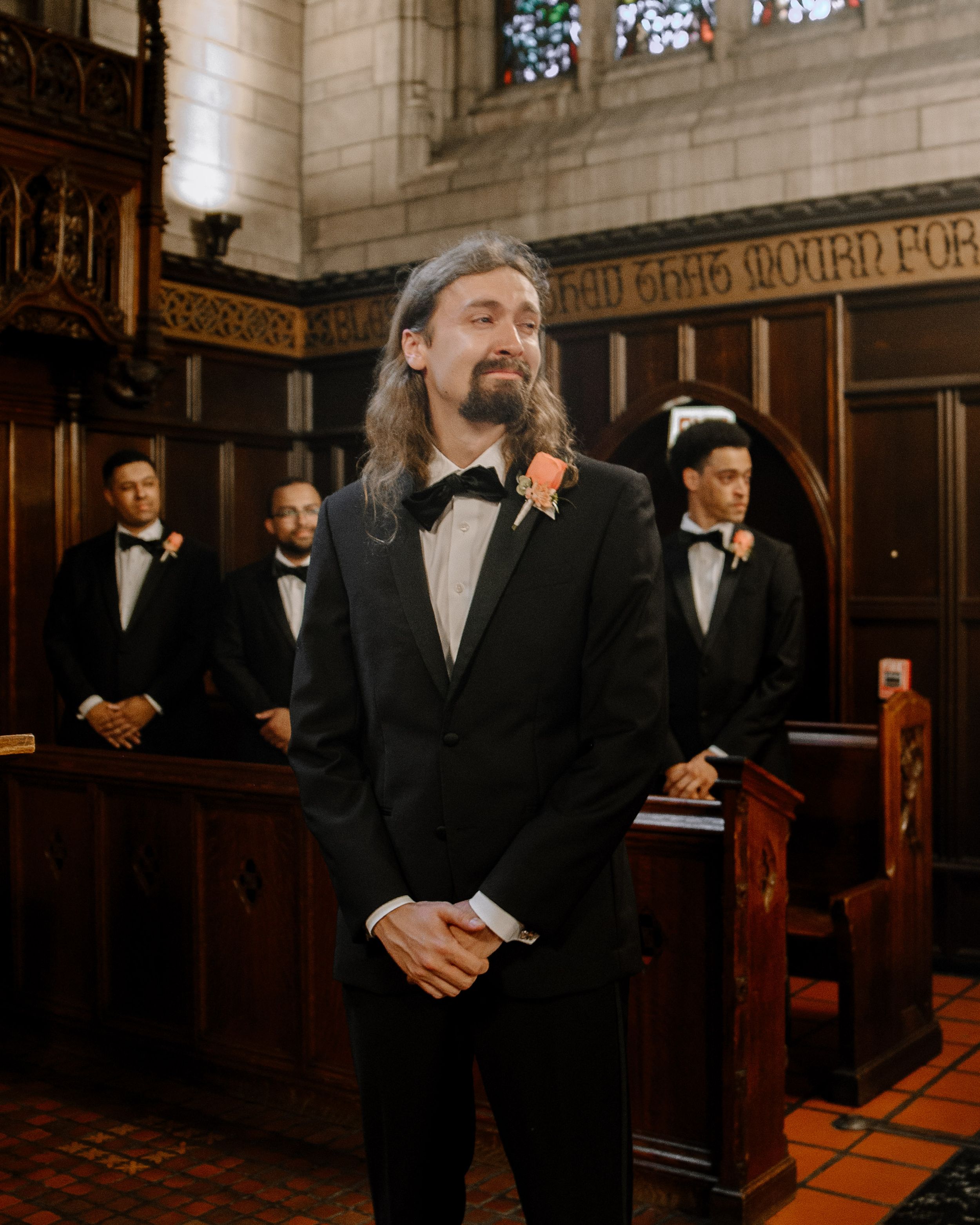 Groomsmen in classic black tuxedos pose in historic church interior with ornate wooden paneling and stained glass windows.