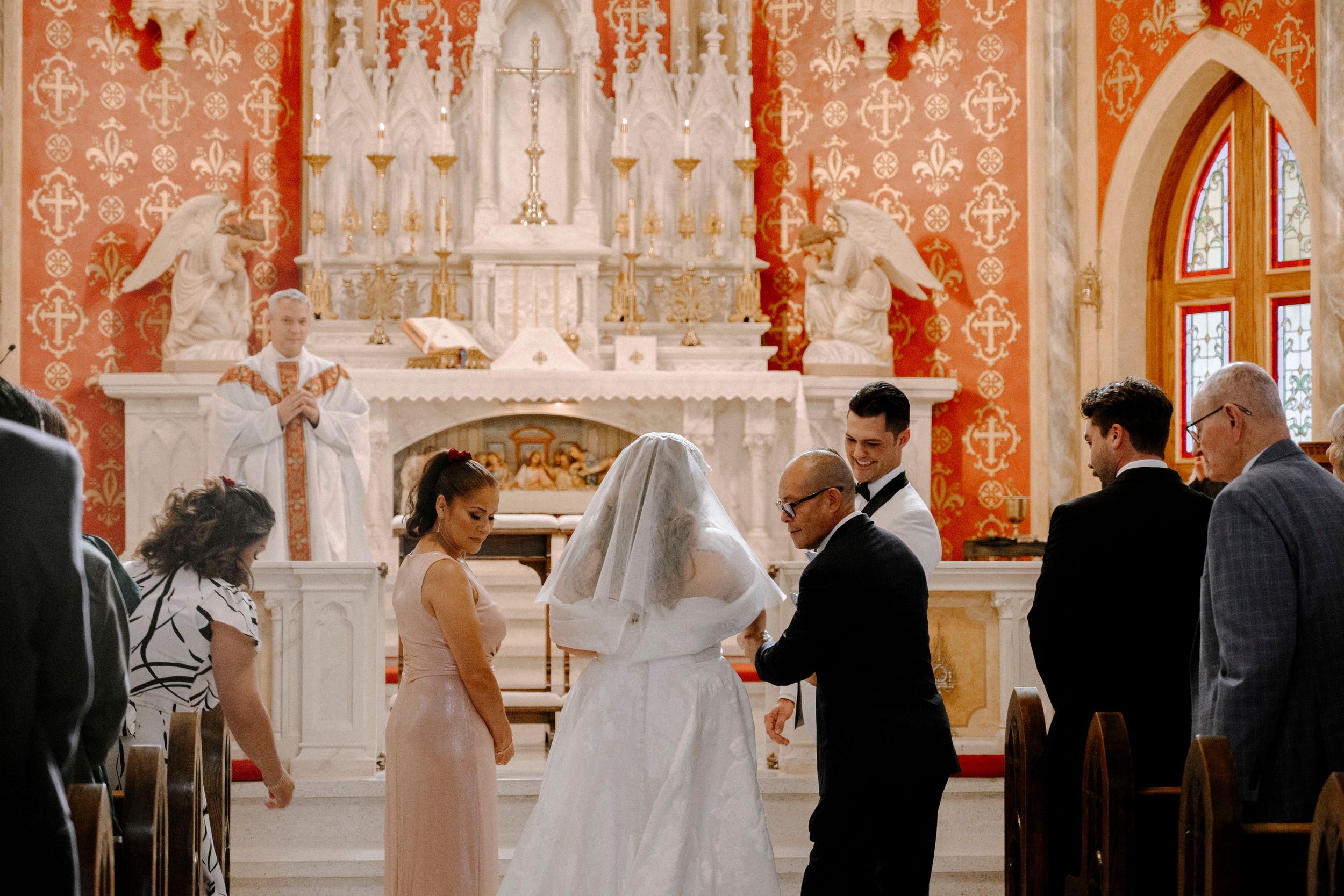 A grand wedding ceremony takes place inside an ornate church with red and gold decor and religious iconography.