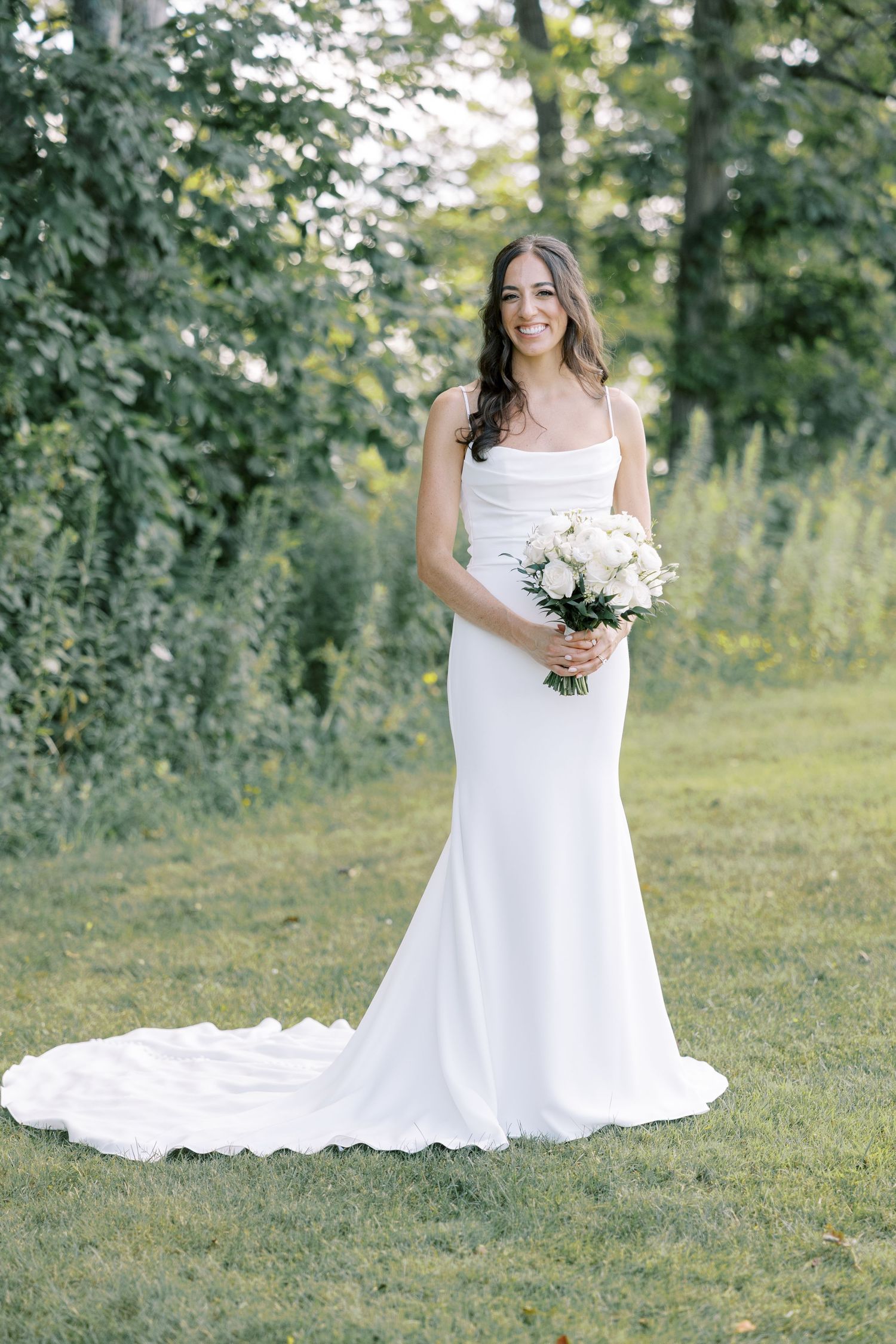 A person in a beautiful white wedding dress poses gracefully in a sunlit garden setting holding a bouquet.