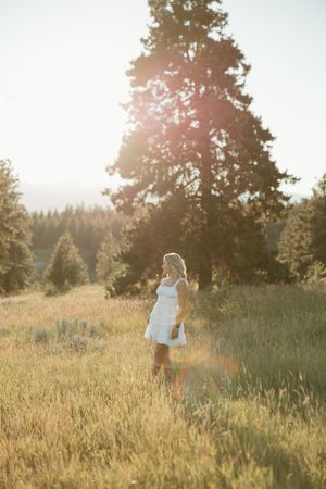 Woman in white dress stands in golden sunlit meadow with evergreen trees at sunset in idyllic nature setting.