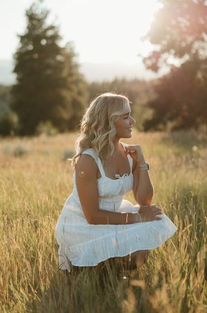 A person in a white sundress crouches in a sunlit meadow during golden hour as tall grass surrounds them.