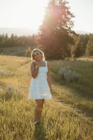 White sundress flows in a golden field at sunset with evergreen trees and mountains in the background.