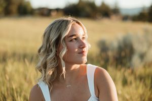 Woman in a white dress sits in a sunlit meadow during golden hour with tall grass swaying in the breeze.