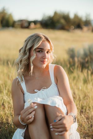 A person in a white dress sits in a sunlit field at golden hour with soft bokeh in the background.