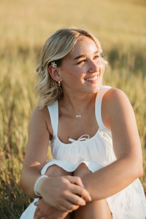 A woman in a white summer dress sits in a golden wheat field at sunset, smiling warmly while bathed in natural light.